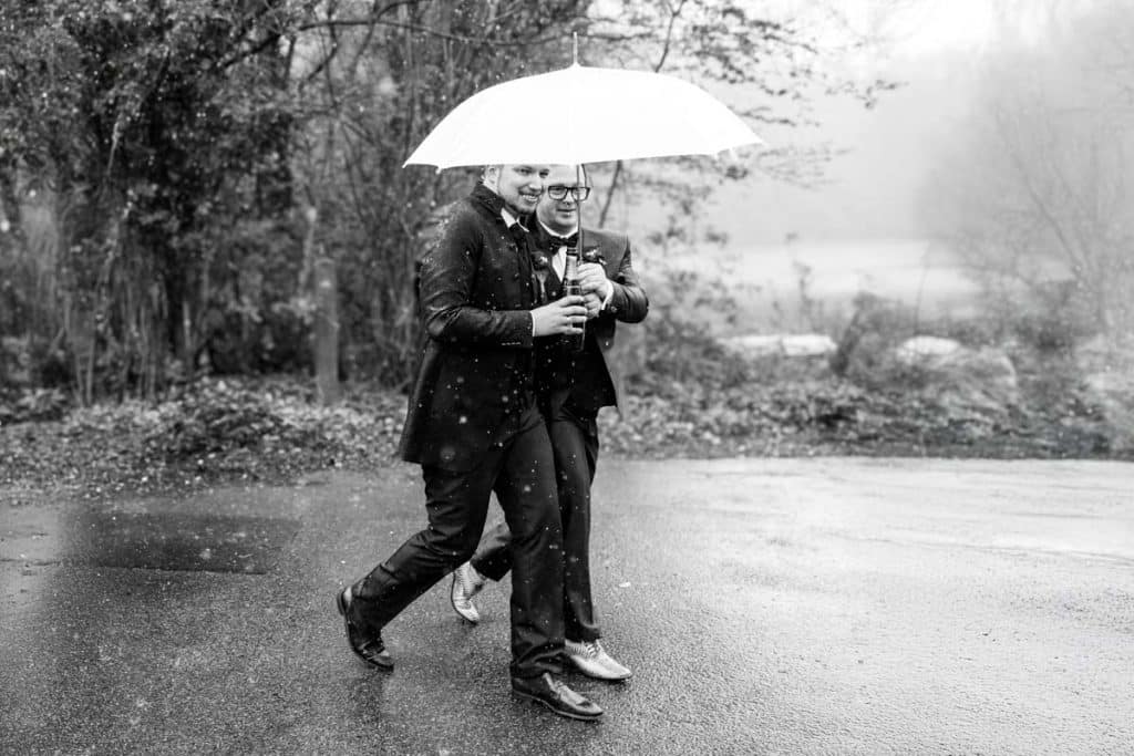 Zwei Bräutigame laufen lachend Arm in Arm unter einem weißen Regenschirm im Regen am Rhein in Duisburg-Alt Walsum. Beide tragen elegante Anzüge mit Fliege. Dieses ausdrucksstarke Schwarz-Weiß-Bild zeigt die Liebe und Freude bei einer gleichgeschlechtlichen Hochzeit, aufgenommen vom Hochzeitsfotograf Duisburg, Wesel, Oberhausen und Dinslaken.