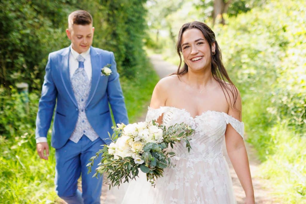 Fröhliche Braut in den Rheinauen Duisburg-Walsum: Die Braut trägt ein schulterfreies Spitzenkleid und hält einen großen Brautstrauß mit weißen Rosen und Eukalyptus, während sie lachend in die Kamera schaut. Ihr Bräutigam im blauen Anzug folgt ihr auf einem Naturweg. Authentische Hochzeitsreportage vom Hochzeitsfotograf Duisburg, Wesel, Oberhausen und Dinslaken