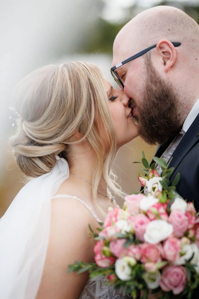 Nahaufnahme eines Brautpaares beim Kuss: Die Braut mit eleganter Hochsteckfrisur und Schleier hält einen farbenfrohen Brautstrauß mit weißen und rosafarbenen Rosen, während sie zärtlich ihren Bräutigam küsst. Der Bräutigam trägt einen dunklen Anzug und Brille