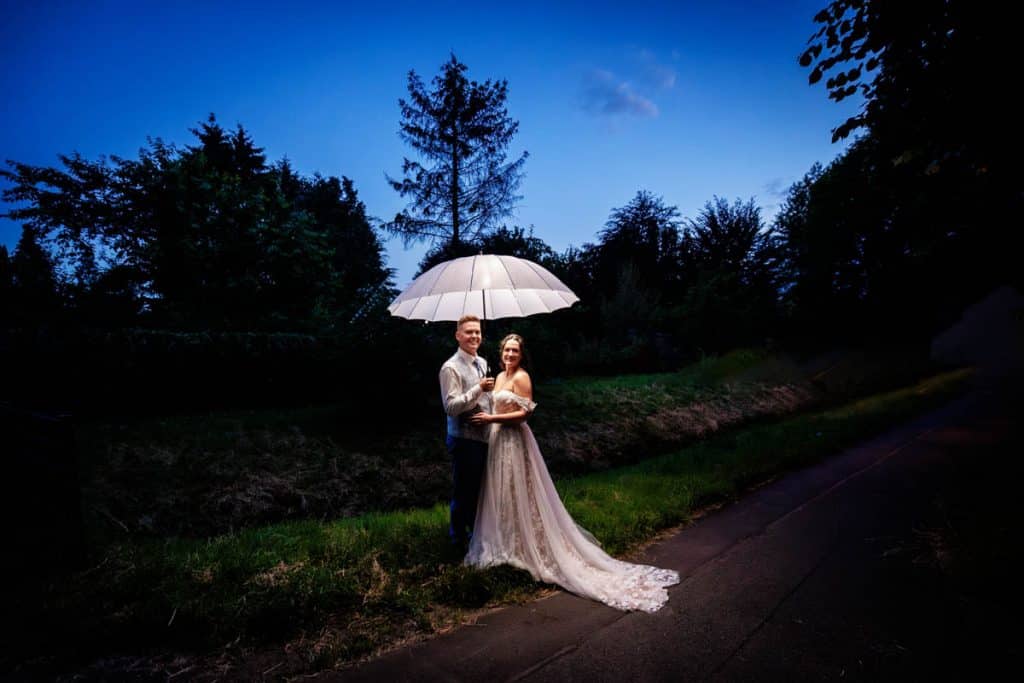 Ein Brautpaar posiert in der Abenddämmerung unter einem großen beleuchteten Regenschirm. Die Braut trägt ein elegantes Spitzenkleid mit Schleppe, der Bräutigam einen hellen Anzug. Das stimmungsvolle Hochzeitsfoto bei Nacht zeigt die romantische Atmosphäre, eingefangen vom Hochzeitsfotograf Duisburg, Wesel und Oberhausen.