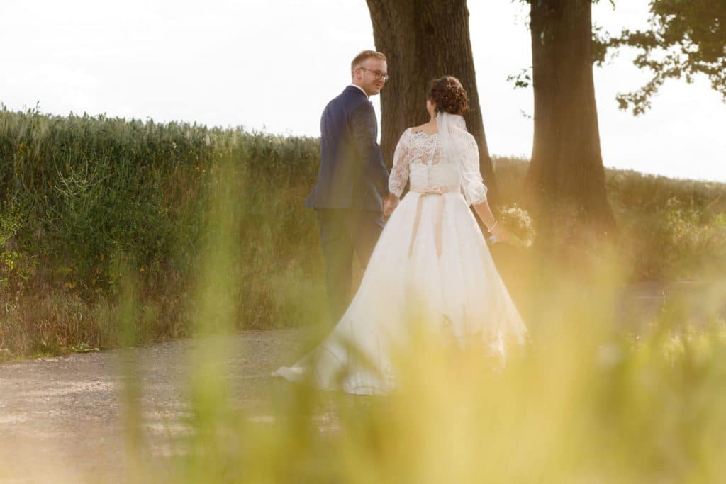 Romantisches Brautpaarshooting im Kurpark in Xanten: Die Braut im eleganten Spitzenkleid und der Bräutigam im festlichen Anzug spazieren Hand in Hand durch die Parkanlage. Sonnenlicht, grüne Bäume und Blumenrabatten schaffen eine idyllische Kulisse. Dieses Foto zeigt die natürliche und moderne Hochzeitsfotografie vom Hochzeitsfotograf Xanten.