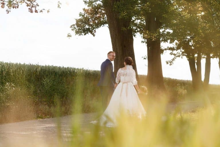 Romantisches Brautpaarshooting im Kurpark in Xanten: Die Braut im eleganten Spitzenkleid und der Bräutigam im festlichen Anzug spazieren Hand in Hand durch die Parkanlage. Sonnenlicht, grüne Bäume und Blumenrabatten schaffen eine idyllische Kulisse. Dieses Foto zeigt die natürliche und moderne Hochzeitsfotografie vom Hochzeitsfotograf Xanten.