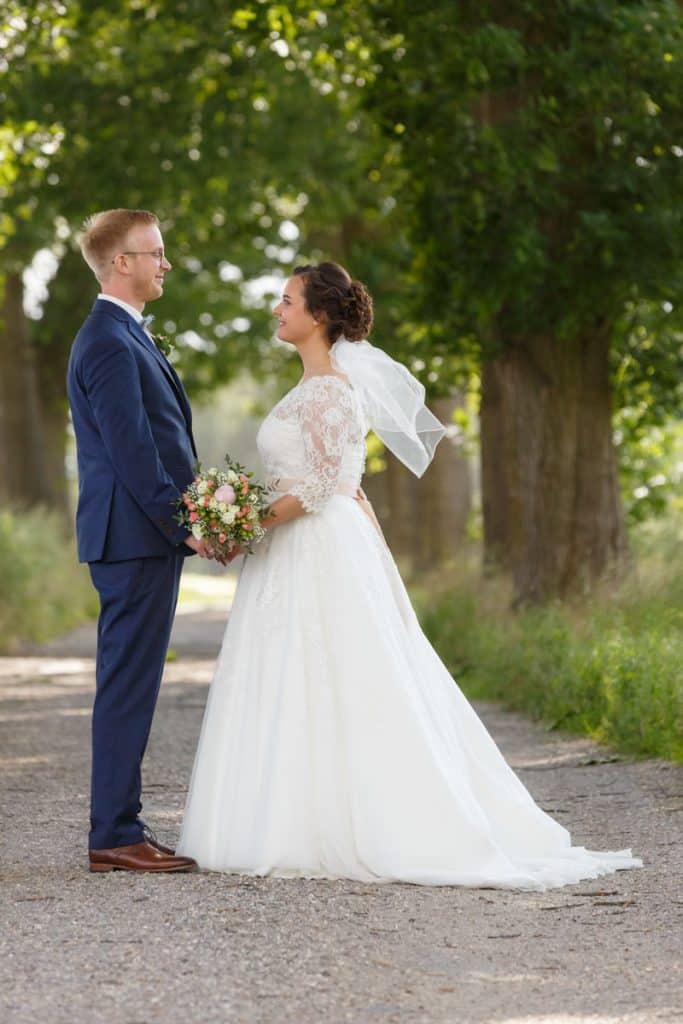 Romantisches Brautpaarshooting im Kurpark in Xanten: Die Braut im eleganten Spitzenkleid und der Bräutigam im festlichen Anzug spazieren Hand in Hand durch die Parkanlage. Sonnenlicht, grüne Bäume und Blumenrabatten schaffen eine idyllische Kulisse. Dieses Foto zeigt die natürliche und moderne Hochzeitsfotografie vom Hochzeitsfotograf Xanten.