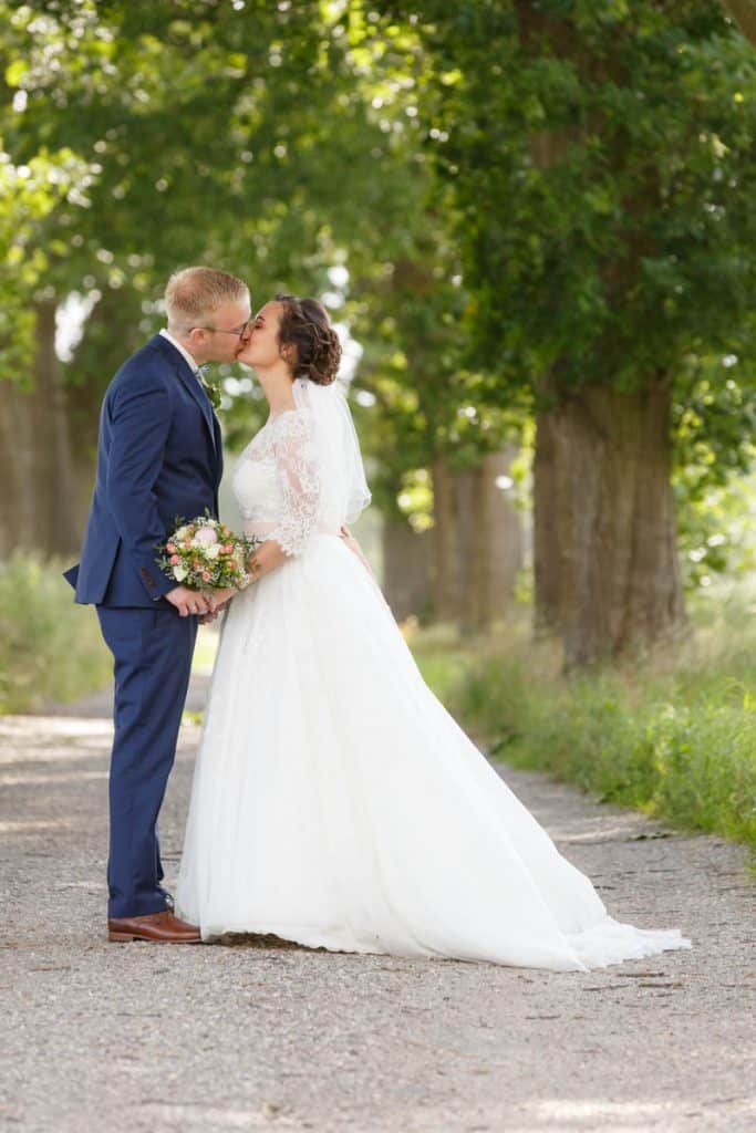 Romantisches Brautpaarshooting im Kurpark in Xanten: Die Braut im eleganten Spitzenkleid und der Bräutigam im festlichen Anzug spazieren Hand in Hand durch die Parkanlage. Sonnenlicht, grüne Bäume und Blumenrabatten schaffen eine idyllische Kulisse. Dieses Foto zeigt die natürliche und moderne Hochzeitsfotografie vom Hochzeitsfotograf Xanten.