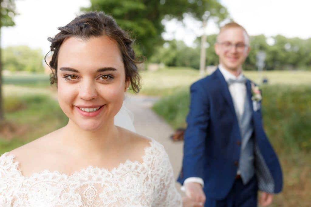 Romantisches Brautpaarshooting im Kurpark in Xanten: Die Braut im eleganten Spitzenkleid und der Bräutigam im festlichen Anzug spazieren Hand in Hand durch die Parkanlage. Sonnenlicht, grüne Bäume und Blumenrabatten schaffen eine idyllische Kulisse. Dieses Foto zeigt die natürliche und moderne Hochzeitsfotografie vom Hochzeitsfotograf Xanten.