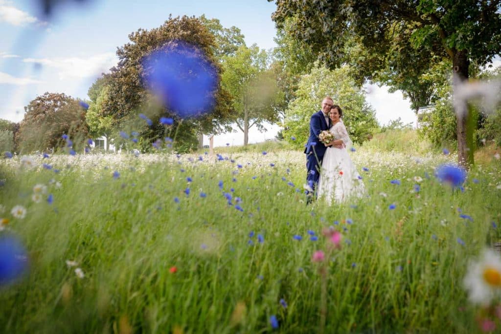 Romantisches Brautpaarshooting im Kurpark in Xanten: Die Braut im eleganten Spitzenkleid und der Bräutigam im festlichen Anzug spazieren Hand in Hand durch die Parkanlage. Sonnenlicht, grüne Bäume und Blumenrabatten schaffen eine idyllische Kulisse. Dieses Foto zeigt die natürliche und moderne Hochzeitsfotografie vom Hochzeitsfotograf Xanten.