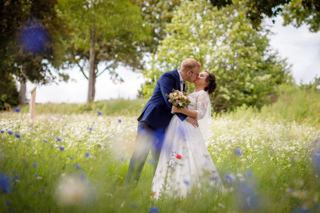 Romantisches Brautpaarshooting im Kurpark in Xanten: Die Braut im eleganten Spitzenkleid und der Bräutigam im festlichen Anzug spazieren Hand in Hand durch die Parkanlage. Sonnenlicht, grüne Bäume und Blumenrabatten schaffen eine idyllische Kulisse. Dieses Foto zeigt die natürliche und moderne Hochzeitsfotografie vom Hochzeitsfotograf Xanten.