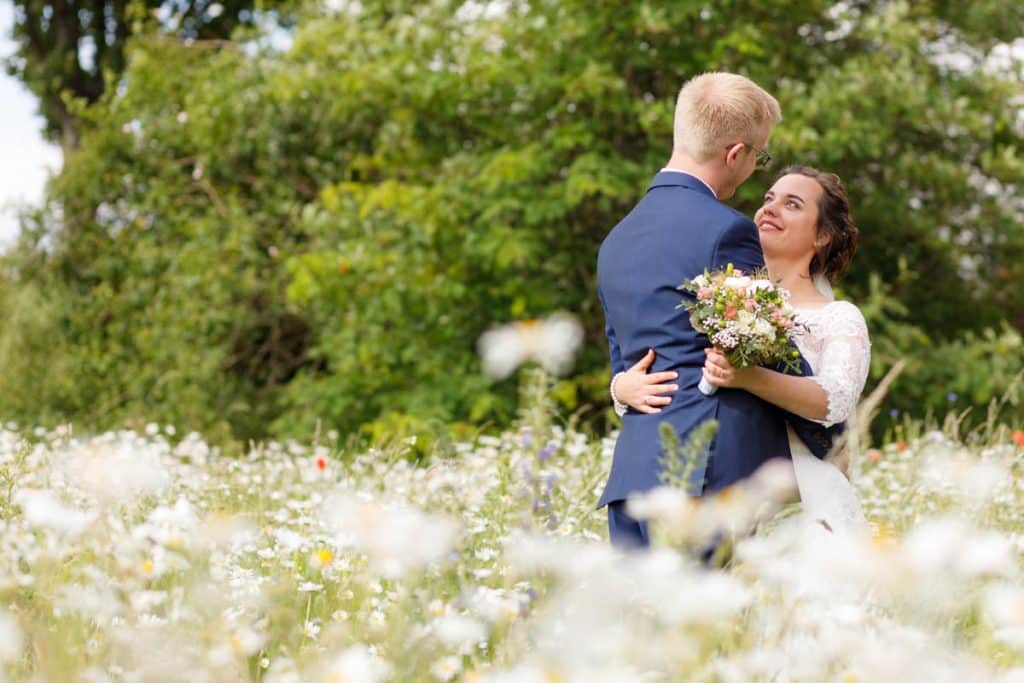 Romantisches Brautpaarshooting im Kurpark in Xanten: Die Braut im eleganten Spitzenkleid und der Bräutigam im festlichen Anzug spazieren Hand in Hand durch die Parkanlage. Sonnenlicht, grüne Bäume und Blumenrabatten schaffen eine idyllische Kulisse. Dieses Foto zeigt die natürliche und moderne Hochzeitsfotografie vom Hochzeitsfotograf Xanten.