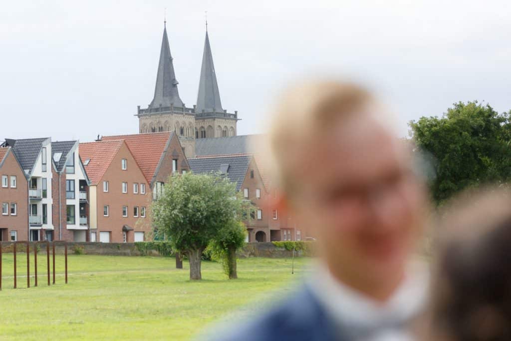 Romantisches Brautpaarshooting im Kurpark in Xanten: Die Braut im eleganten Spitzenkleid und der Bräutigam im festlichen Anzug spazieren Hand in Hand durch die Parkanlage. Sonnenlicht, grüne Bäume und Blumenrabatten schaffen eine idyllische Kulisse. Dieses Foto zeigt die natürliche und moderne Hochzeitsfotografie vom Hochzeitsfotograf Xanten.