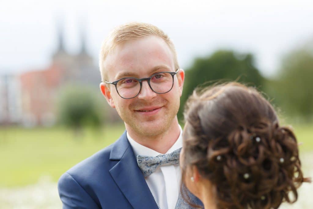 Romantisches Brautpaarshooting im Kurpark in Xanten: Die Braut im eleganten Spitzenkleid und der Bräutigam im festlichen Anzug spazieren Hand in Hand durch die Parkanlage. Sonnenlicht, grüne Bäume und Blumenrabatten schaffen eine idyllische Kulisse. Dieses Foto zeigt die natürliche und moderne Hochzeitsfotografie vom Hochzeitsfotograf Xanten.