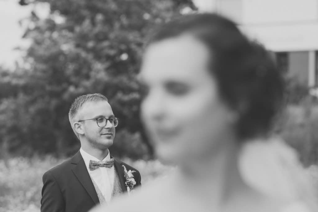 Romantisches Brautpaarshooting im Kurpark in Xanten: Die Braut im eleganten Spitzenkleid und der Bräutigam im festlichen Anzug spazieren Hand in Hand durch die Parkanlage. Sonnenlicht, grüne Bäume und Blumenrabatten schaffen eine idyllische Kulisse. Dieses Foto zeigt die natürliche und moderne Hochzeitsfotografie vom Hochzeitsfotograf Xanten.