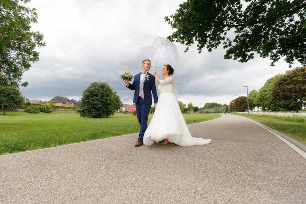 Romantisches Brautpaarshooting im Kurpark in Xanten: Die Braut im eleganten Spitzenkleid und der Bräutigam im festlichen Anzug spazieren Hand in Hand durch die Parkanlage. Sonnenlicht, grüne Bäume und Blumenrabatten schaffen eine idyllische Kulisse. Dieses Foto zeigt die natürliche und moderne Hochzeitsfotografie vom Hochzeitsfotograf Xanten.
