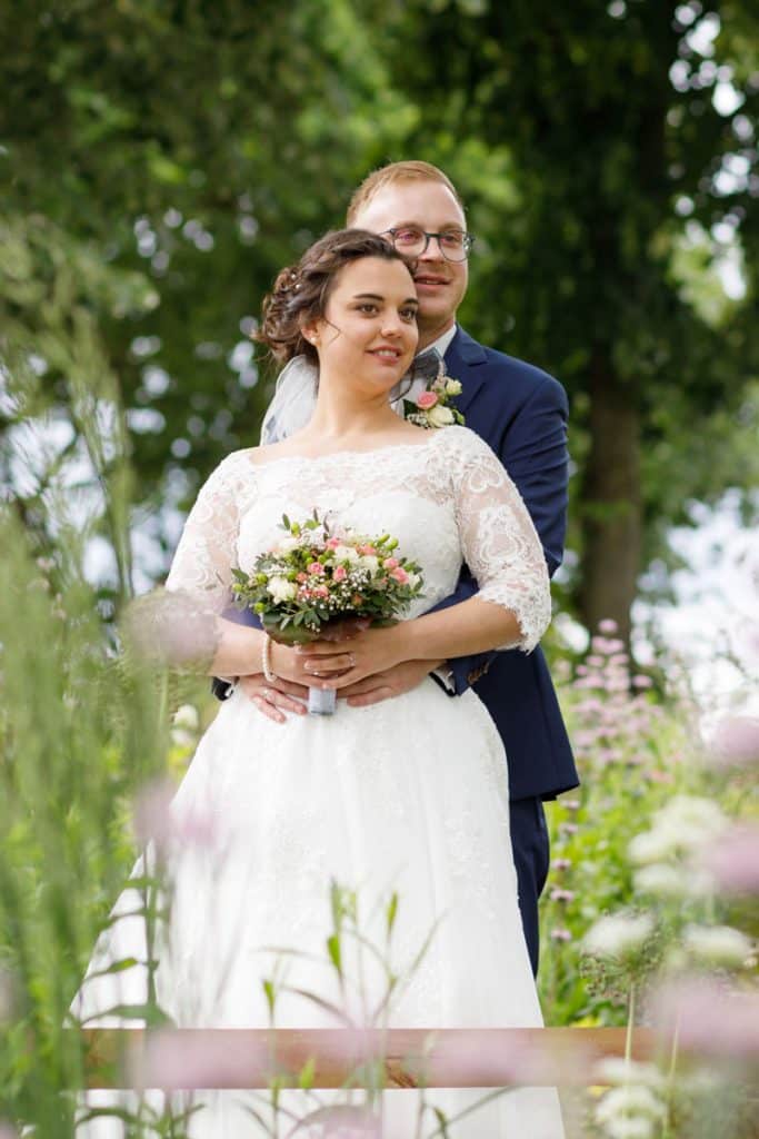 Romantisches Brautpaarshooting im Kurpark in Xanten: Die Braut im eleganten Spitzenkleid und der Bräutigam im festlichen Anzug spazieren Hand in Hand durch die Parkanlage. Sonnenlicht, grüne Bäume und Blumenrabatten schaffen eine idyllische Kulisse. Dieses Foto zeigt die natürliche und moderne Hochzeitsfotografie vom Hochzeitsfotograf Xanten.