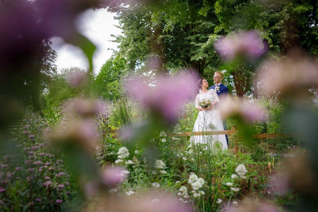 Romantisches Brautpaarshooting im Kurpark in Xanten: Die Braut im eleganten Spitzenkleid und der Bräutigam im festlichen Anzug spazieren Hand in Hand durch die Parkanlage. Sonnenlicht, grüne Bäume und Blumenrabatten schaffen eine idyllische Kulisse. Dieses Foto zeigt die natürliche und moderne Hochzeitsfotografie vom Hochzeitsfotograf Xanten.