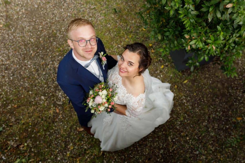 Romantisches Brautpaarshooting im Kurpark in Xanten: Die Braut im eleganten Spitzenkleid und der Bräutigam im festlichen Anzug spazieren Hand in Hand durch die Parkanlage. Sonnenlicht, grüne Bäume und Blumenrabatten schaffen eine idyllische Kulisse. Dieses Foto zeigt die natürliche und moderne Hochzeitsfotografie vom Hochzeitsfotograf Xanten.