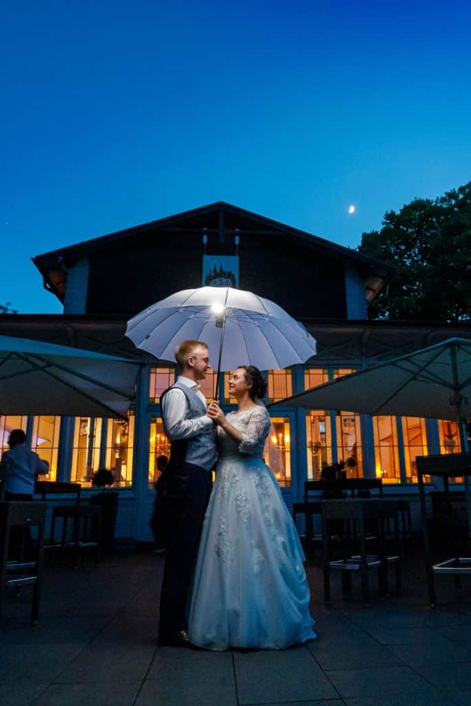 Romantisches Brautpaarshooting im Kurpark in Xanten: Die Braut im eleganten Spitzenkleid und der Bräutigam im festlichen Anzug spazieren Hand in Hand durch die Parkanlage. Sonnenlicht, grüne Bäume und Blumenrabatten schaffen eine idyllische Kulisse. Dieses Foto zeigt die natürliche und moderne Hochzeitsfotografie vom Hochzeitsfotograf Xanten.