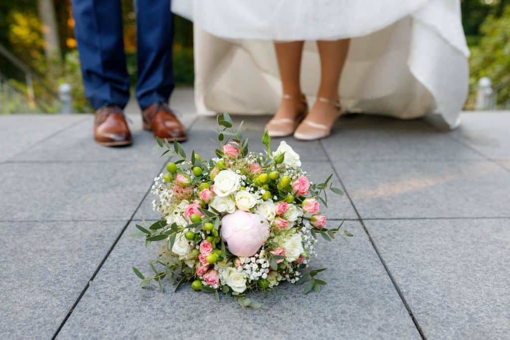 Romantisches Brautpaarshooting im Kurpark in Xanten: Die Braut im eleganten Spitzenkleid und der Bräutigam im festlichen Anzug spazieren Hand in Hand durch die Parkanlage. Sonnenlicht, grüne Bäume und Blumenrabatten schaffen eine idyllische Kulisse. Dieses Foto zeigt die natürliche und moderne Hochzeitsfotografie vom Hochzeitsfotograf Xanten.