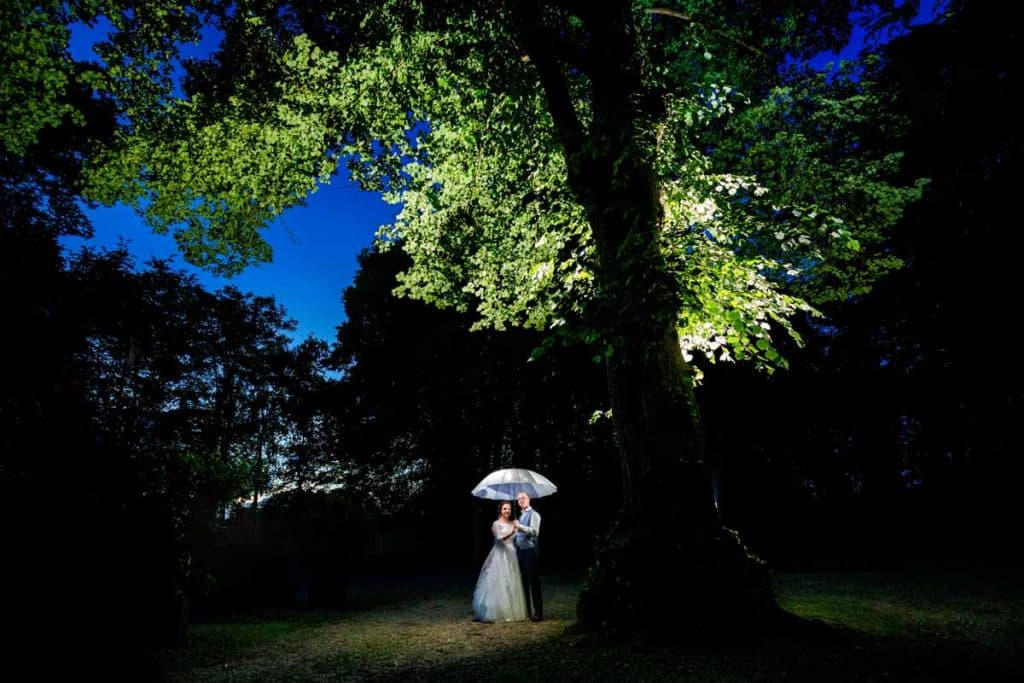 Romantisches Brautpaarshooting im Kurpark in Xanten: Die Braut im eleganten Spitzenkleid und der Bräutigam im festlichen Anzug spazieren Hand in Hand durch die Parkanlage. Sonnenlicht, grüne Bäume und Blumenrabatten schaffen eine idyllische Kulisse. Dieses Foto zeigt die natürliche und moderne Hochzeitsfotografie vom Hochzeitsfotograf Xanten.