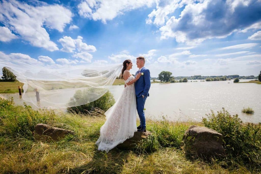 Romantisches Brautpaar am Rhein in Duisburg-Walsum: Die Braut im Spitzenkleid mit langem, wehenden Schleier steht auf einem Stein, während der Bräutigam im blauen Anzug liebevoll in ihre Augen blickt. Im Hintergrund die weitläufigen Rheinauen mit blauem Himmel und weißen Wolken. Kreative und emotionale Hochzeitsfotografie vom Hochzeitsfotograf Duisburg, Wesel, Dinslaken und Oberhausen