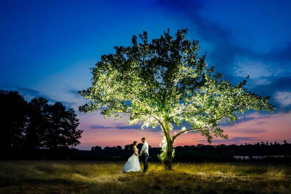 Romantisches Hochzeitsfoto eines Brautpaares unter einem hell erleuchteten Baum in der Abenddämmerung. Der Himmel zeigt kräftige Farben des Sonnenuntergangs in Blau und Rosa. Die Braut trägt ein elegantes Brautkleid, der Bräutigam einen hellen Anzug. Ein einzigartiges Paarshooting in der Natur, festgehalten vom Hochzeitsfotograf Duisburg und NRW.