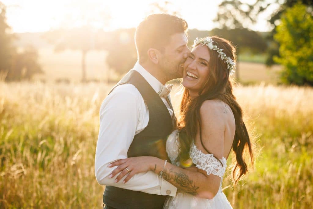 Emotionales Hochzeitsfoto eines Brautpaares im goldenen Licht der Abendsonne: Die Braut lacht herzlich, während der Bräutigam sie liebevoll umarmt und küsst. Ein romantischer Moment, aufgenommen vom Hochzeitsfotograf Wesel Landgut am Hochwald Sonsbeck