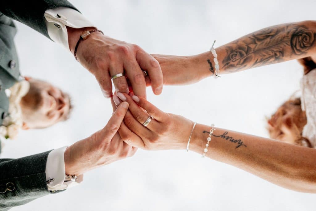 Kreatives Hochzeitsfoto aus der Froschperspektive: Braut und Bräutigam halten ihre frisch angesteckten Eheringe in die Kamera. Sichtbar sind liebevolle Details wie Tattoos, Armbänder und elegant manikürte Fingernägel. Dieses emotionale Detailfoto wurde vom Hochzeitsfotograf Landgut am Hochwald Sonsbeck