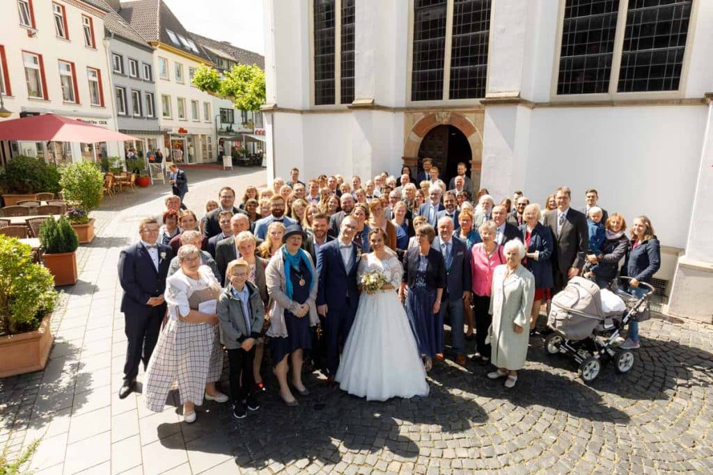 Fröhliche Gruppenfotos nach der Hochzeit in Xanten: Das Brautpaar posiert mit Familie und Freunden vor der Kirche. Lächelnde Gesichter, elegante Outfits und die festliche Stimmung machen die Gruppenfotos zu einer bleibenden Erinnerung. Fotografiert vom Hochzeitsfotograf Xanten.
