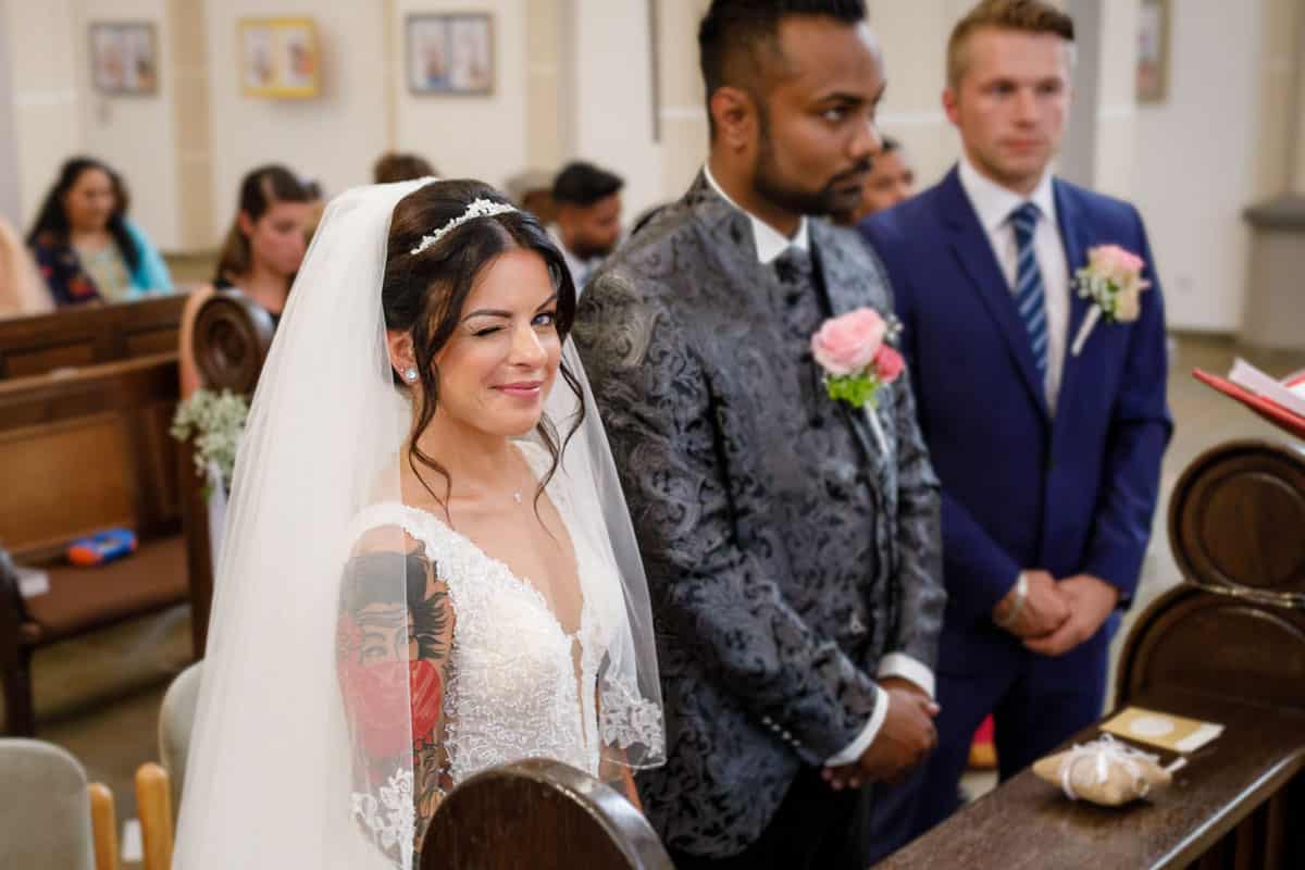 Emotionaler Moment während der kirchlichen Trauung: Die Braut im weißen Spitzenkleid mit Tattoo und Schleier steht lächelnd am Altar und zwinkert verschmitzt in die Kamera. Neben ihr der Bräutigam im festlichen Anzug mit Blumenanstecker. Hinter dem Paar sitzen die Gäste in der Kirche. Ein authentisches Hochzeitsfoto, das die Freude und Leichtigkeit des Augenblicks einfängt – fotografiert vom Hochzeitsfotograf Duisburg, Wesel und Oberhausen.