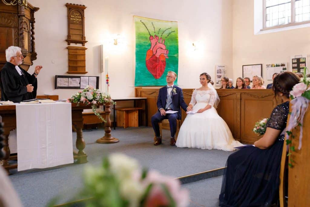 Emotionaler Moment bei der kirchlichen Trauung in der Evangelischen Kirche am Markt in Xanten: Das Brautpaar steht gemeinsam am Altar, die Braut im weißen Spitzenkleid mit Schleier, der Bräutigam im eleganten Anzug. Gäste verfolgen die Zeremonie aufmerksam in der festlich geschmückten Kirche. Dieses Foto zeigt die authentische Hochzeitsreportage vom Hochzeitsfotograf Xanten.
