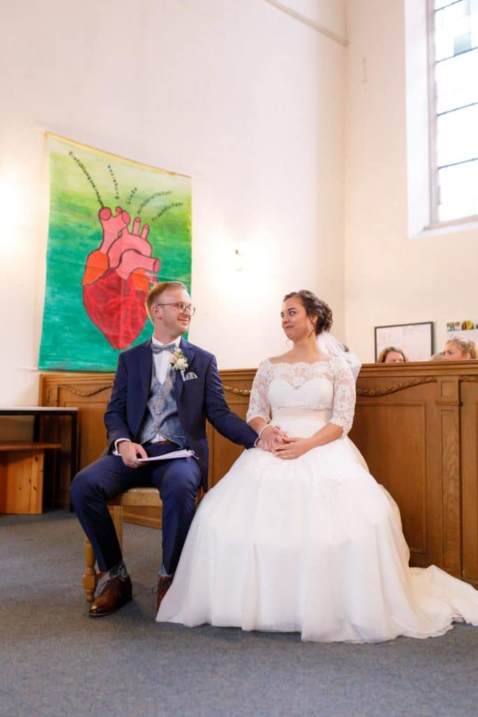 Emotionaler Moment bei der kirchlichen Trauung in der Evangelischen Kirche am Markt in Xanten: Das Brautpaar steht gemeinsam am Altar, die Braut im weißen Spitzenkleid mit Schleier, der Bräutigam im eleganten Anzug. Gäste verfolgen die Zeremonie aufmerksam in der festlich geschmückten Kirche. Dieses Foto zeigt die authentische Hochzeitsreportage vom Hochzeitsfotograf Xanten.