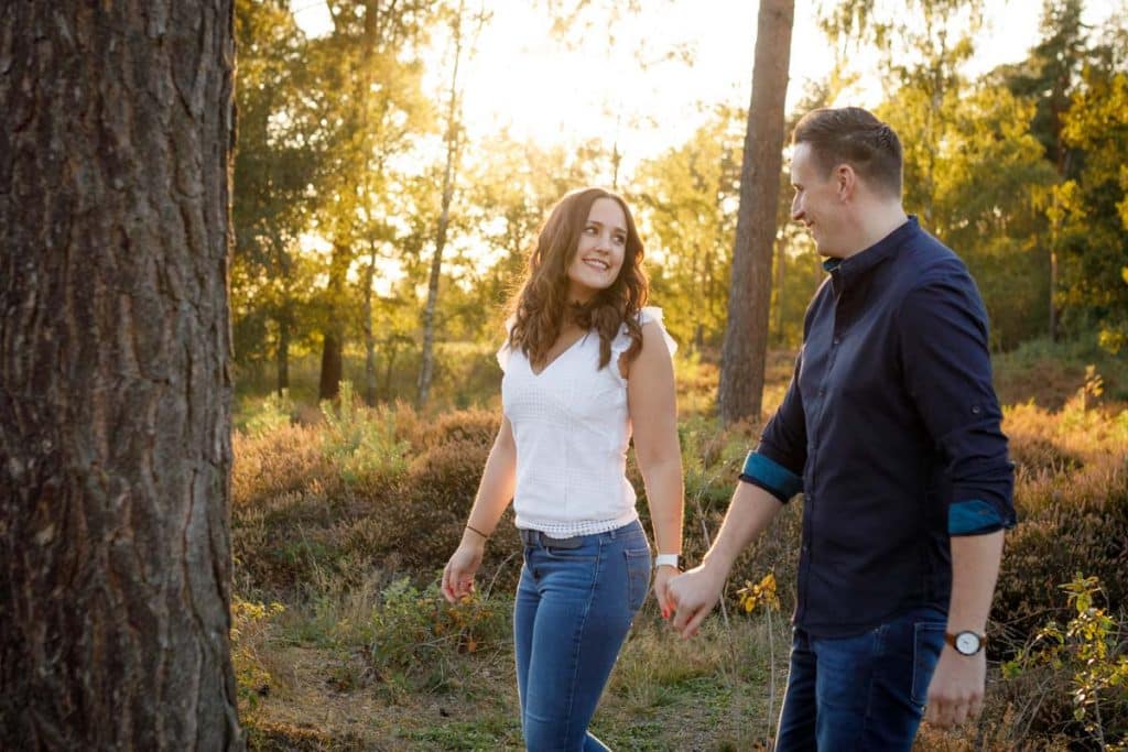 Ein Paar läuft lachend Hand in Hand durch einen sonnendurchfluteten Waldweg. Beide tragen Freizeitkleidung, die natürliche Umgebung und das goldene Licht des Sonnenuntergangs verstärken die Leichtigkeit und Liebe dieses Verlobungsshootings. Fotografiert vom Hochzeitsfotograf Duisburg, Wesel und Oberhausen.