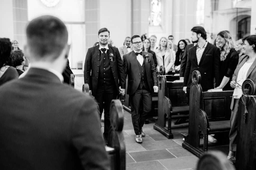 Zwei Bräutigame schreiten Hand in Hand den Mittelgang einer Kirche in Duisburg-Alt Walsum entlang, begleitet von lächelnden Gästen. Beide tragen dunkle Anzüge mit Fliege. Bewegende Hochzeitsreportage in Schwarz-Weiß, fotografiert vom Hochzeitsfotograf Duisburg, Wesel, Oberhausen und Dinslaken.