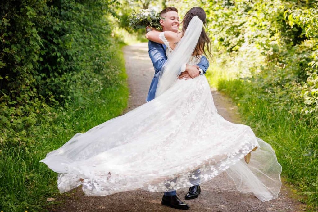 Glücklicher Moment eines Brautpaares in den Rheinauen Duisburg-Walsum: Der Bräutigam hebt die Braut lachend hoch und dreht sie, während ihr langes Spitzenkleid und der Schleier elegant durch die Luft fliegen. Lebendige und emotionale Hochzeitsfotografie vom Hochzeitsfotograf Duisburg, Wesel, Oberhausen und Dinslaken.