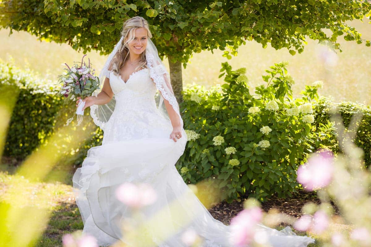 Eine glückliche Braut in einem eleganten Spitzenkleid posiert unter einem Baum. Mit einem Blumenstrauß in der Hand und einem Lächeln im Gesicht entsteht ein romantisches Hochzeitsfoto in der Natur.