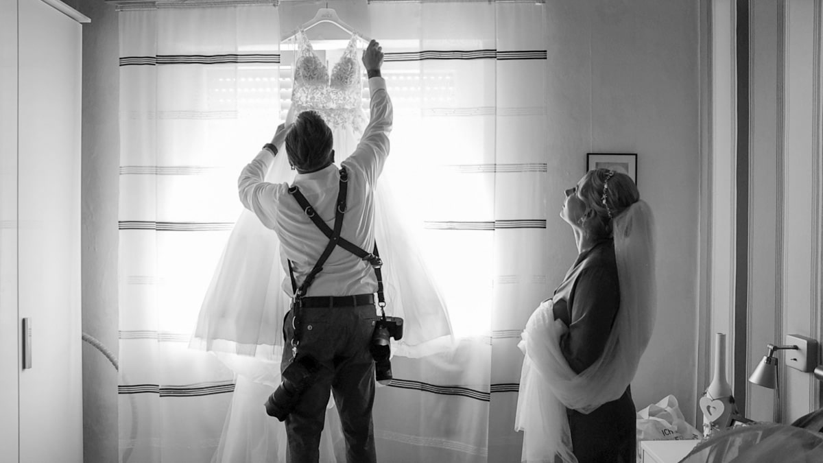 Schwarz-Weiß-Aufnahme eines Hochzeitsfotografen, der ein Brautkleid am Fenster arrangiert, während die Braut daneben steht und zuschaut. Authentischer Einblick hinter die Kulissen einer Hochzeitsreportage.
