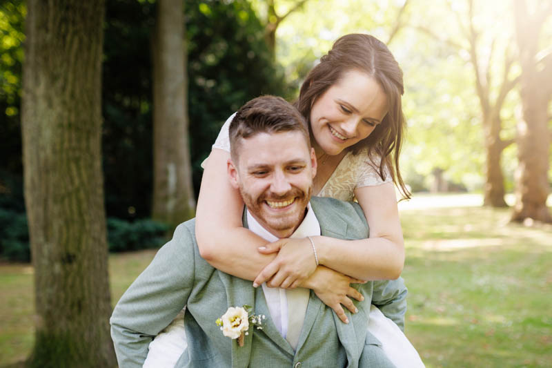 Brautpaarshooting im Grünen: Ein lachendes Brautpaar genießt den Hochzeitstag, die Braut sitzt auf dem Rücken des Bräutigams, beide strahlen und wirken gelöst und fröhlich. Das natürliche Paarfoto wurde vom Hochzeitsfotograf Duisburg, Wesel, Oberhausen und Dinslaken aufgenommen und zeigt die entspannte und liebevolle Stimmung am Hochzeitstag.